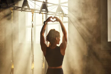 Fotobehang I love active life. Back view of young athletic woman in black sport clothing making heart shape with her hands while training in dark gym © Friends Stock