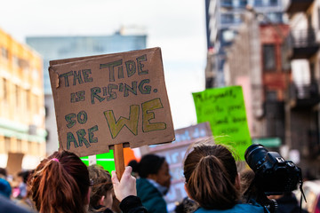 Activists unite for climate change. A closeup view of a cardboard sign reading the tide is rising, so are we, above a gathering of environmental demonstrators on a city street.