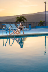 Young lady poses sitting at the border of a swimming pool during summer vacation in dusk. 