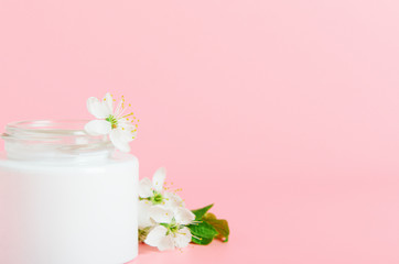 Face cream in white jar on a pink background with white flowers. Concept natural cosmetics, organic beauty. Copy space.