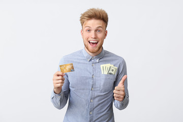 Happy young man with credit card and dollars showing thumb-up gesture on white background