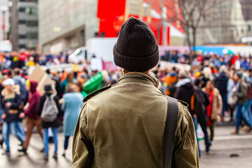 Person watches environmental protest. A young guy wearing a green coat is viewed from behind, watching environmentalists march in the city center of Montreal, Canada