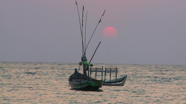 Medium low-angle still shot of a ball of deep orange sun, setting in dull grey sea clouds , above Burmese anchored fishing boats, Ngapali Beach, Myanmar