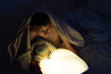 Adorable boy reads book before sleeping in bed at home. Happy childhood.
