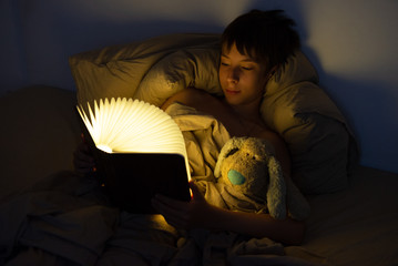 Adorable boy reads book before sleeping in bed at home with his old toy. Happy childhood.