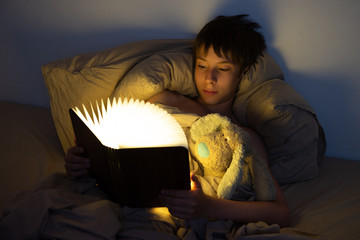 Adorable boy reads book before sleeping in bed at home with his old toy. Happy childhood.