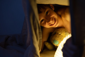 Adorable boy reads book before sleeping in bed at home with his old toy. Happy childhood.