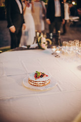 Beautiful simple wedding cake with fruits and champagne glasses on table at wedding reception outdoors in the evening. Wedding celebration and catering in soft candle light in night.