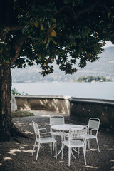 Beautiful provence white table and chairs under tree on shore of Lago Maggiore, Stresa city, Italy. Relaxing on pier on Lago Maggiore. Summer vacation in Europe