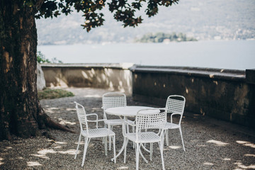 Fototapeta premium Beautiful provence white table and chairs under tree on shore of Lago Maggiore, Stresa city, Italy. Relaxing on pier on Lago Maggiore. Summer vacation in Europe
