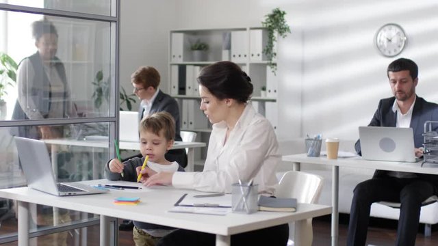 Businesswoman sitting at desk in the office, talking to kid and working with documents while little son drawing on paper with colored pencils