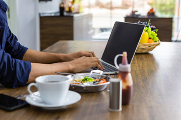 Closeup of young asian man working laptop eating breakfast drinking coffee on wooden table in kitchen room