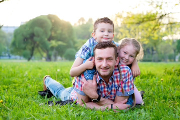 Naklejka na ściany i meble Happy and smile family in park, boy and girl have fun with Dad on green grass and looking at camera. Happy loving family concept