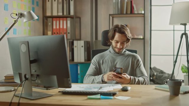 Handsome Long Haired Entrepreneur Sitting at His Desk in the Office Works on Desktop Computer, Working with Documents, Graphs. Smilingly Uses Smartphone, Social Media App, Writing Emails, Messaging