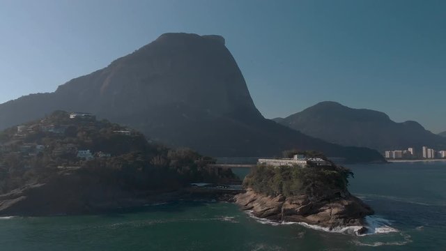 Rotational aerial pan showing the Joatinga beach on a hazy backlit day with the Gavea mountain behind revealing the wider well known cityscape of Rio de Janeiro in the background