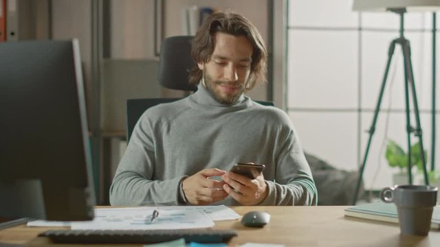 Handsome Long Haired Entrepreneur Sitting at His Desk in the Office Works on Desktop Computer, Working with Documents, Graphs. Uses Smartphone, Social Media App, Writing Emails, Messaging