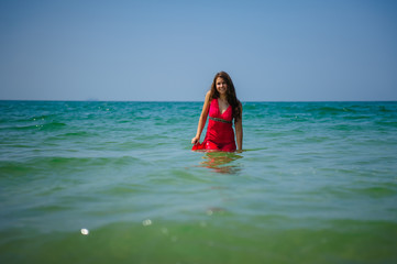 Young sexy long-haired brunette in red beach dress stands in the turquoise water of the ocean on a hot day. Beautiful smiling woman posing in the sea during vacation.