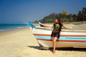 Beautiful sexy brunette on tropical sandy beach near wooden boat on blue sea background and clear sky on hot sunny day. A girl with long hair in a transparent tunic stands near colorful boat.