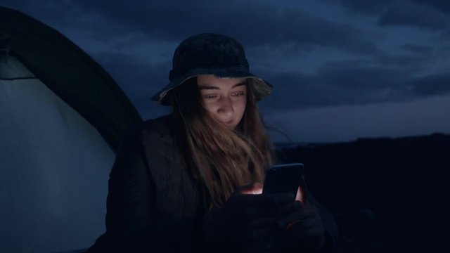 Happy and stoked young adventurous camping woman sits in dawn or night darkness next to tent at camp ground, ssmartphone screen lits her face, uses outdoor application, chat or text