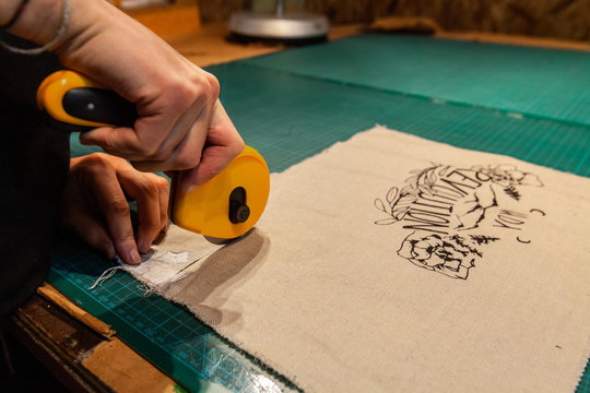 Fashion designer is cutting patches. A close-up view on the hands of a quilter using a rotary cutter on a patch of beige fabric with floral design. Textile preparation in clothes making.