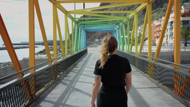 Young female traveler or tourist explores new city or town, walk on colorful bright rainbow bridge at empty boardwalk at sunny summer beach. Happy explorer woman, student exchange concept