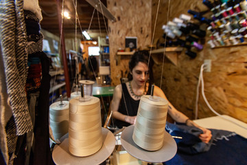 Fashion designer using vintage overlock. A creative clothes designer is viewed sitting at workstation. Young woman uses overlock machine with white thread spools inside workshop.