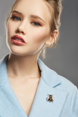 Portrait of girl with tied back fair hair, wearing sky blue coat with bright brooch in view of black fly on lapel. The woman is tilting head and looking at camera, posing against the gray background.