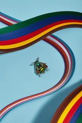 Closeup shot of breastpin in view of green fly with wings and contrasting stripes, isolated asymmetrically on blue background in midst of multicolored wavy placed ribbons. Voguish women's accessory.