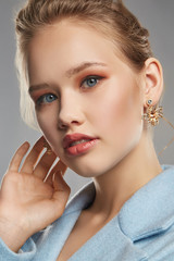 Fototapeta na wymiar Closeup shot of blonde lady with plush lips, wearing sky blue coat and massive golden earring, adorned with pendant in view of bee with crystals. The girl is looking at camera on gray background.