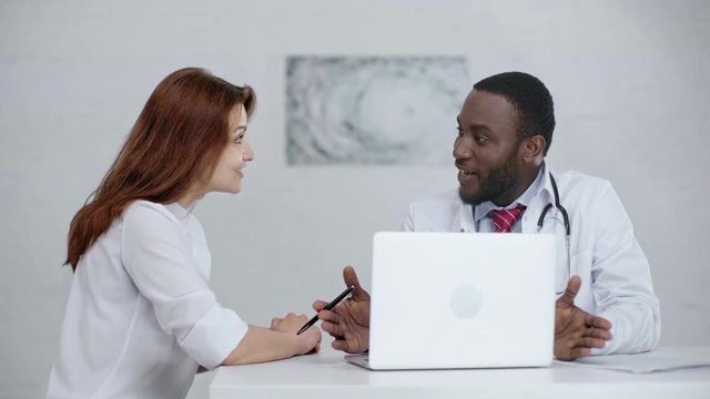two multicultural doctors talking and smiling while sitting near laptop 