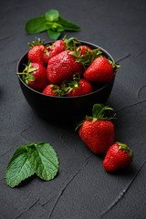 Heap of fresh strawberries and mint leaves in black bowl on dark background. top view