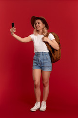 Naklejka premium Portrait of a girl with curly blond hair dressed in a sundwon hat and white t-shirt standing on a red background making selfie with mobile phone. Model travels with orange backpack.