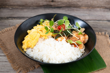 Rice topped with stir-fried shrimp and basil, and fried eggs in white dish.