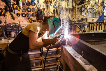 Fototapeta Tradesman operates MIG welder indoors. A side profile view of a blacksmith concentrating on welding two steel beams together with a MIG welder. Hot sparks are created during the joining process. obraz