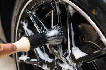 A male worker washes a black car with a special brush for cast wheels and scrubs the surface to shine in a vehicle detailing workshop. Auto service industry.
