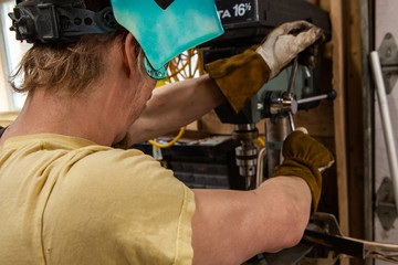 Metalworker operates drill press in shed. View over the shoulder of a metalworker wearing personal protective equipment whilst operating a pedestal drill inside a workshop.