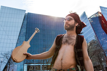 Shirtless man with headband and merlin. A sacred man is seen shirtless in the city center, front view of hairy chest on Caucasian male, holds a small wooden guitar and seeks inspiration.