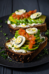 Avocado sandwich on dark rye bread made with fresh sliced avocado, chopped tomatoes, eggs, cream cheese. black wood background