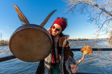 Boho guy holds mystical objects by river. A spiritual man holds traditional objects on a riverbank during sunset. Viewed from in front, he holds leather drum to camera and look off to the side.