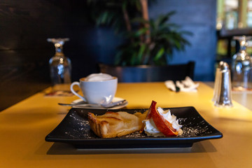 Apple tart served with cream in bistro. A closeup view on a freshly served apple pie with whipped cream on a table inside a restaurant. Blurred tableware is seen in background with copy space above.