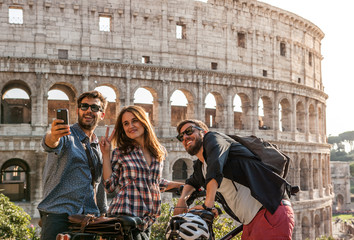 Three happy young friends tourists with bikes at Colosseum in Rome taking pictures and selfies with smartphone