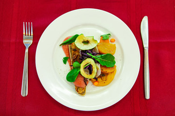 Freshly prepared vegan appetizer. A top down view of a healthy avocado salad served on a white ceramic plate, with cutlery against a red table, vegan appetizer neatly presented.