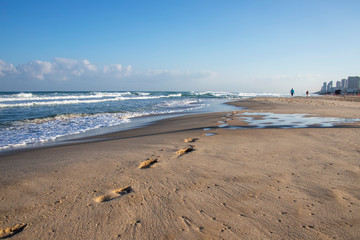 Footprints on the sand of a Mediterranean beach. Foamy waves and buildings on the horizon.