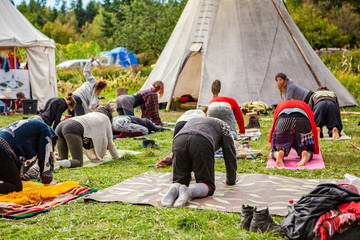 Fusion of cultural & modern music event. A group of people are seen doing yoga stretches near tipi tents in a festival camping area, mats are laid out on grass as group are on all fours. © Valmedia