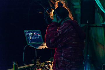 Fusion of cultural & modern music event. A young music deejay is viewed from behind by night, using headphones and electronic equipment to play beats at an eclectic festival outdoors
