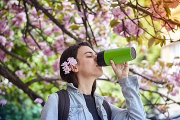 Young woman drinking from green bottle on the background of blooming sakura