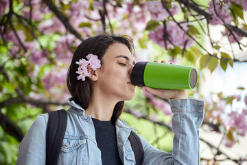 Young woman drinking from green bottle on the background of blooming sakura