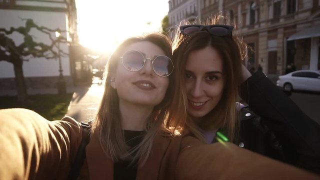 Caucasian fashion women in sunglasses posing for selfie and laughing. Looking at the camera, front view. Lifestyle portrait on street city and sunlight on background