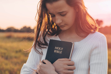Christian teenage girl holds bible in her hands. Reading the Holy Bible in a field during beautiful...