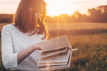 Christian teenage girl holds book in her hands. Reading the book in a field during beautiful sunset.  The girl sitting on a grass, reading a book. Rest and reading